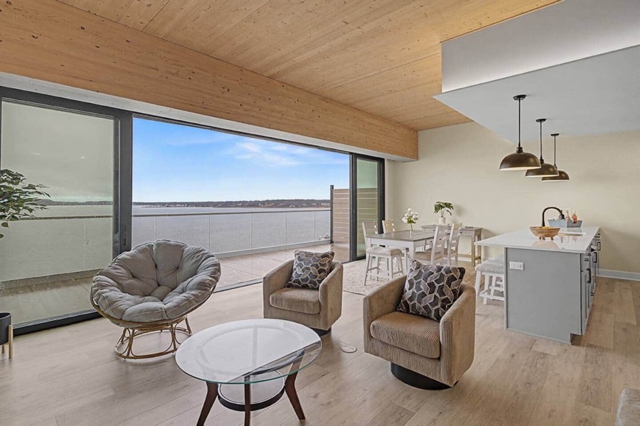 Bright corner living space of Residence 602 — a papasan chair and swivel armchairs beside a wall of glass sliders opening to a Muskegon Lake balcony