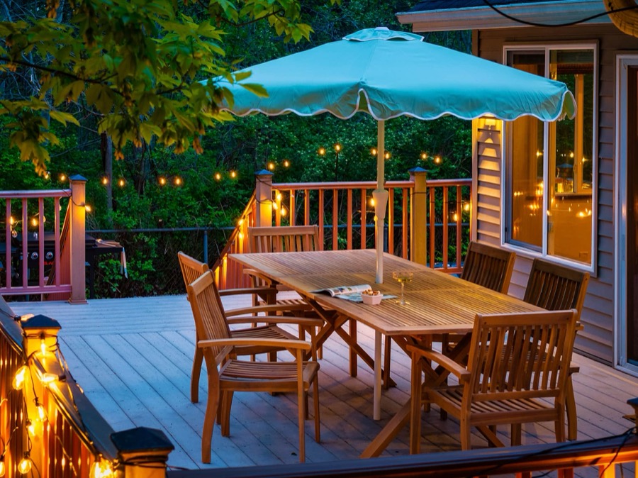 Teak outdoor dining table for six under a scalloped aqua umbrella on the string-lit back deck at dusk