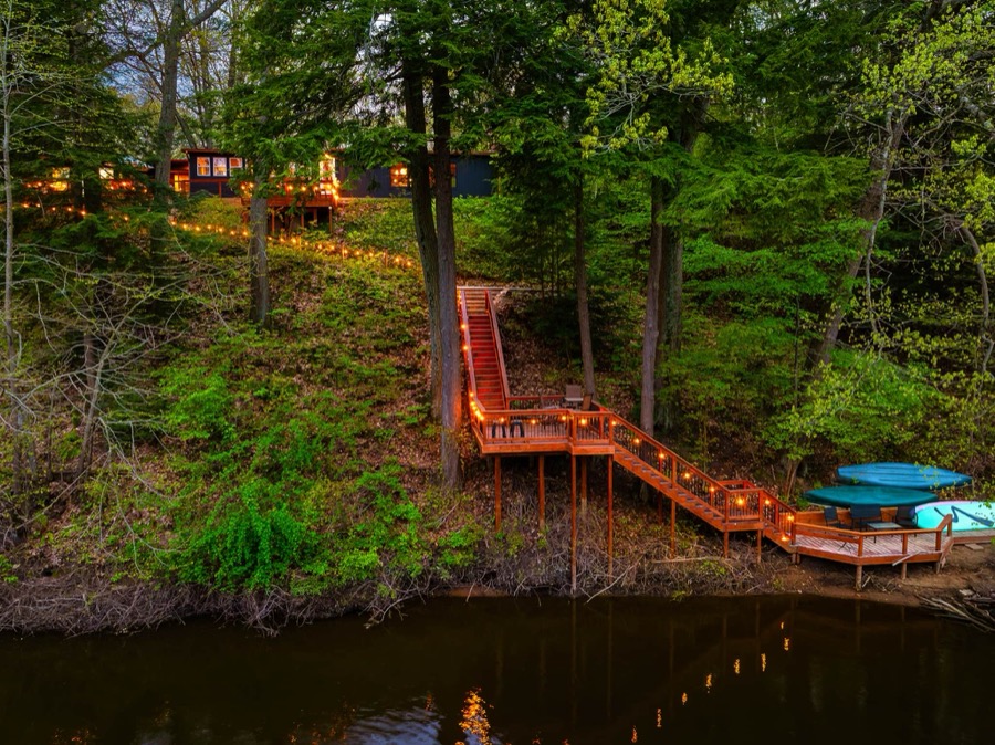 Aerial view of the lantern-lit stairway winding down the wooded bluff to the water-level deck and private dock, kayaks and paddleboards waiting at the Kalamazoo River
