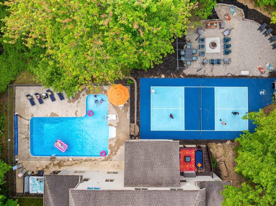 Aerial view of the Swimming in Amenities estate in Holland — L-shaped inground pool with loungers, private blue pickleball court, and a gravel fire-pit lounge tucked into the trees