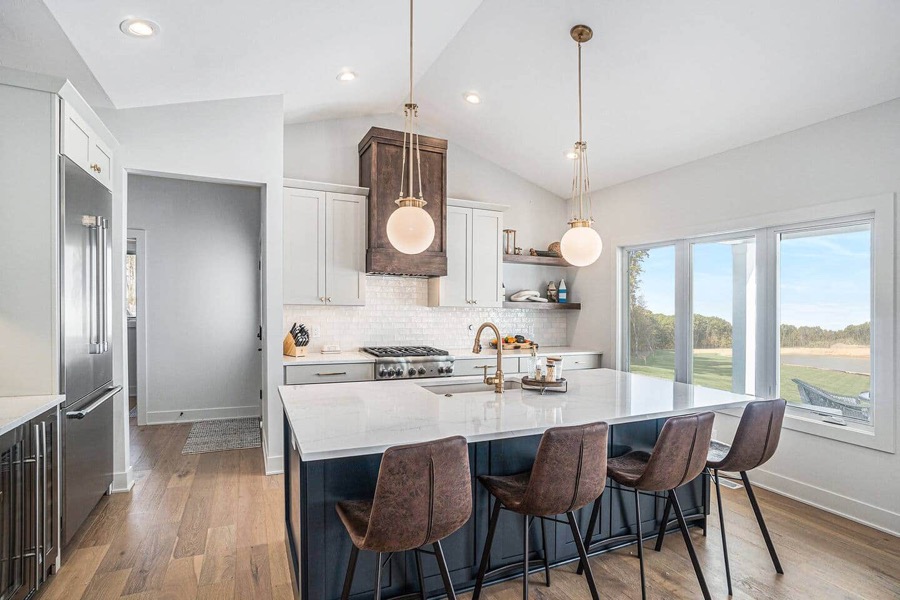 The white chef's kitchen: brass globe pendants, a long marble island with leather stools, and windows to the lake