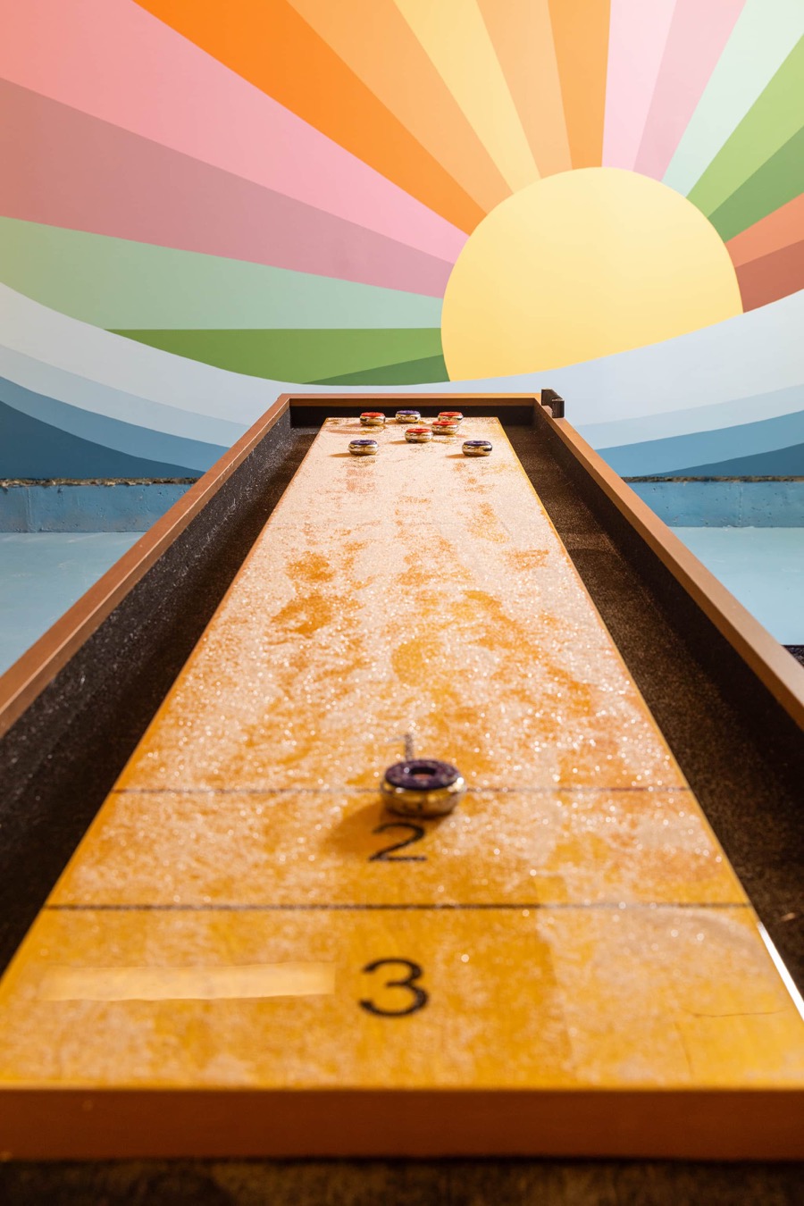 Shuffleboard table in the Swimming in the Sunset game room, set against a hand-painted rainbow sunburst mural