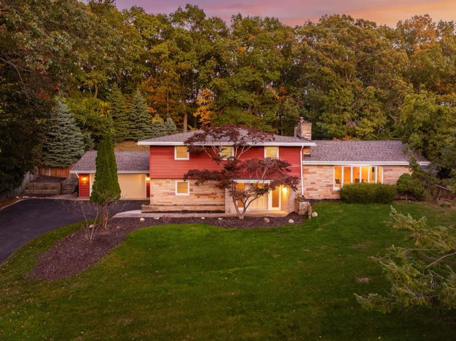 The red-sided tri-level home glowing at dusk under a pink sky, set back on its big wooded lawn